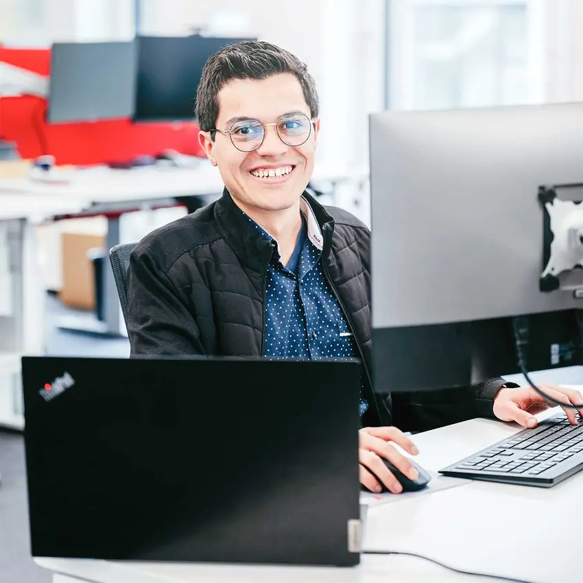 Ein junger Mann mit Brille sitzt an einem Schreibtisch in einem modernen Büro, arbeitet an zwei Monitoren und lächelt in die Kamera.